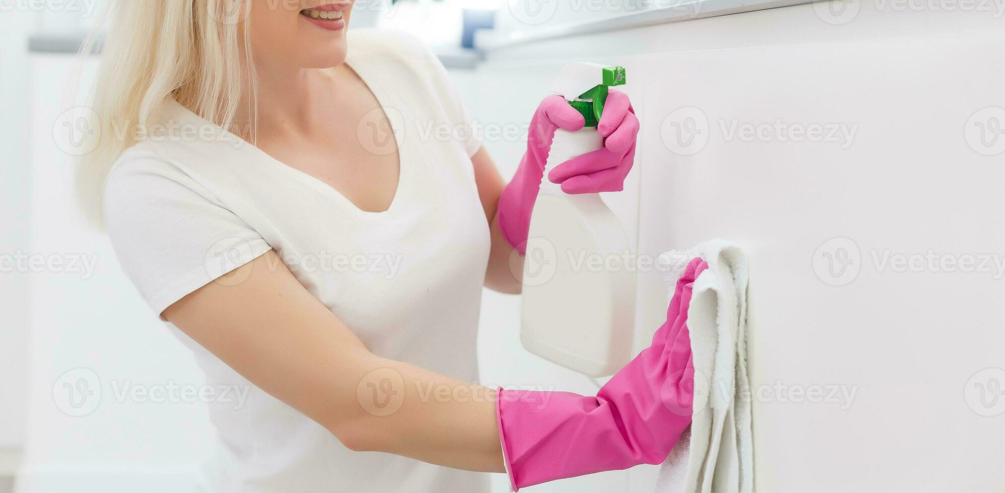 Woman in the kitchen is smiling and wiping dust using a spray and a duster while cleaning her house, close-up photo