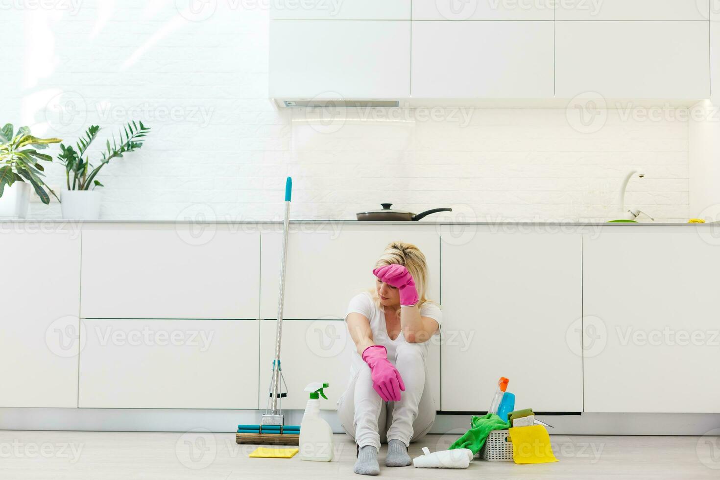 Tired young cleaning lady resting in the kitchen after house keeping.Sitting on kitchen countertop and holding mop and spray bottle photo