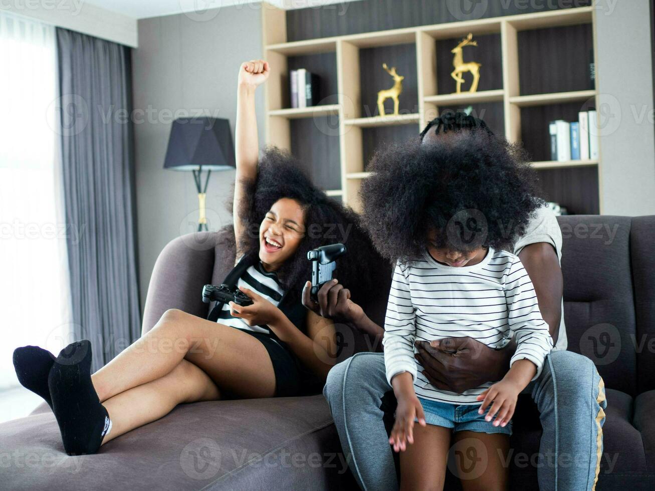 son girl kid female woman lady person people human afro hair black dark skin pretty beautiful sit sofa bed indoor home house apartment hotel condominium together play game joystick laugh cheerful dad photo