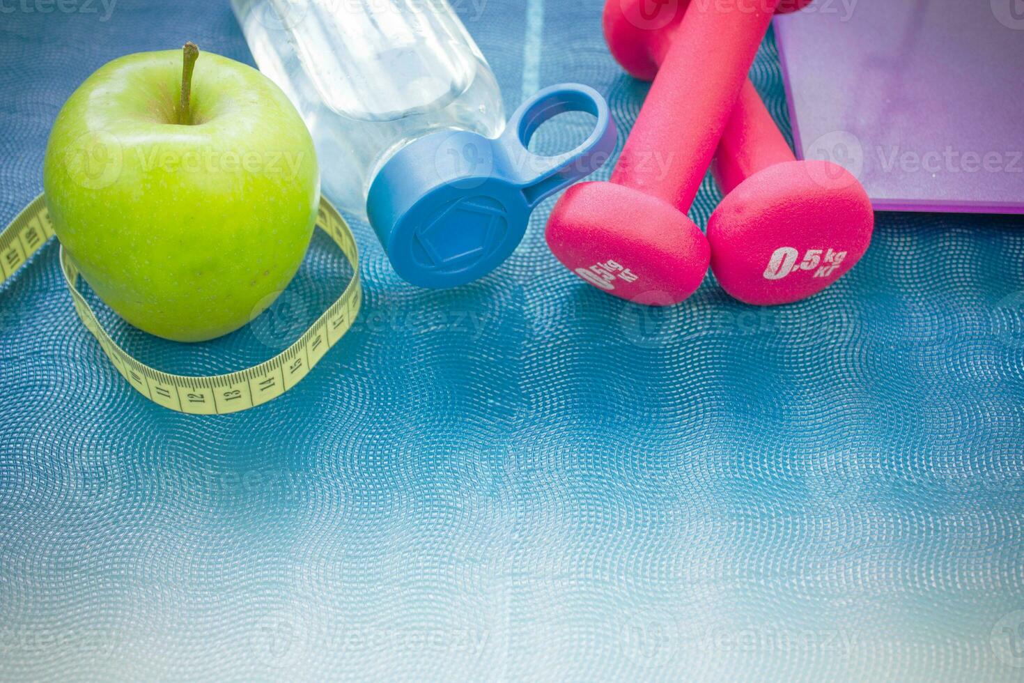 Two red dumbbells, an apple, a tape measure, a bottle of water and weight scales on a yoga mat photo