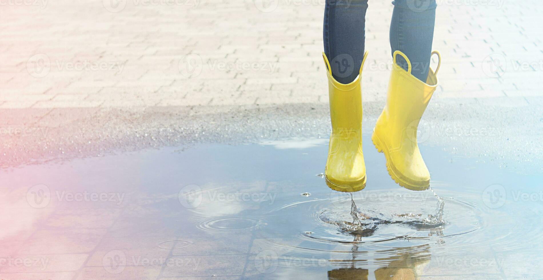 Happy child in yellow wellington boot jumps in a puddle of water after rain. Cheerful baby on a summer sunny day. Outdoor games. Copy space photo