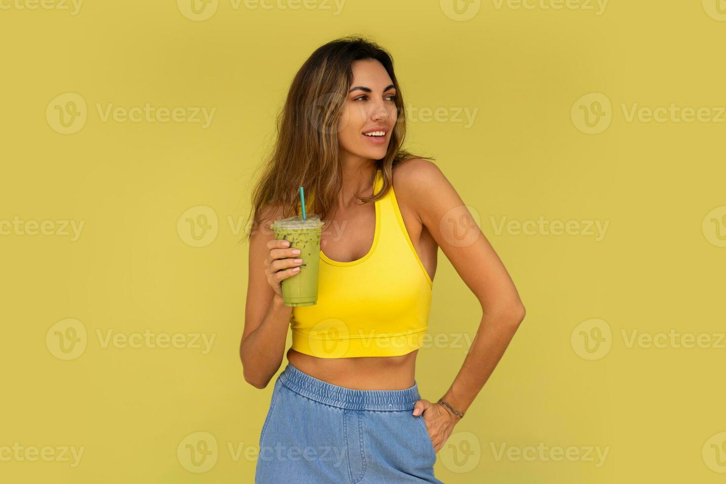 estudio foto de bonito morena mujer en juguetón atuendo hoding bebida y posando en amarillo antecedentes. Perfecto Delgado cuerpo.