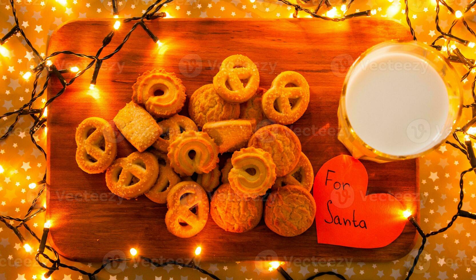 Wooden board with yellow cookies, glass of milk, paper heart For Santa and surrounded with Christmas lights photo