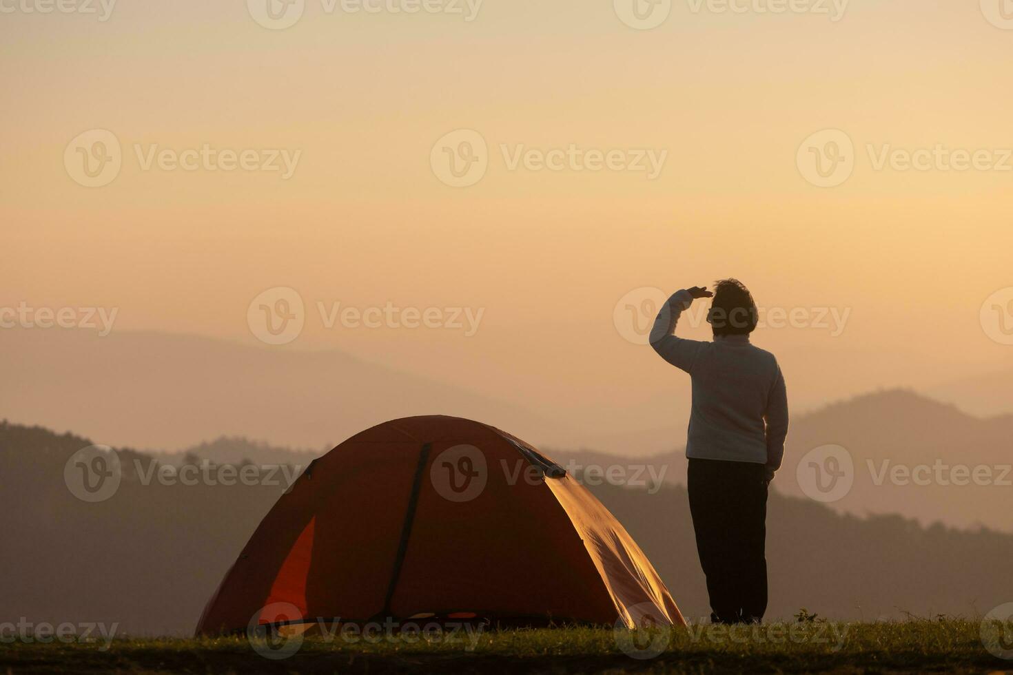 Traveller is standing by the tent during overnight camping while looking at the beautiful scenic sunset over the mountain for outdoor adventure vacation travel photo