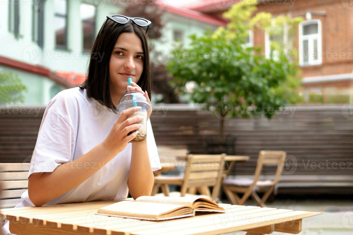 un linda morena niña es sentado a un mesa en un verano restaurante con un enfriamiento bebida foto