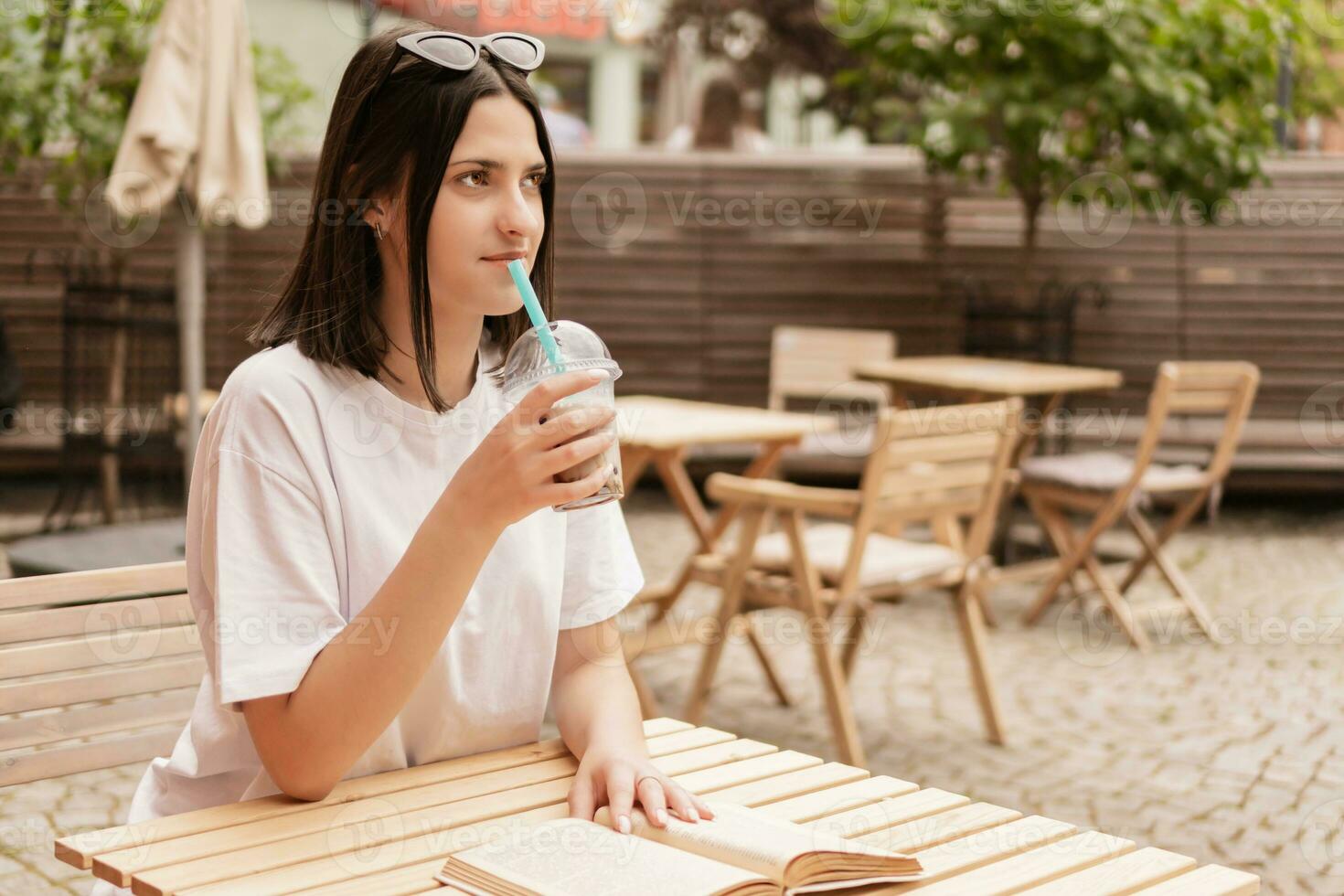 A young girl sits at a table in a summer cafe, drinks a drink through a straw and reads a book. photo