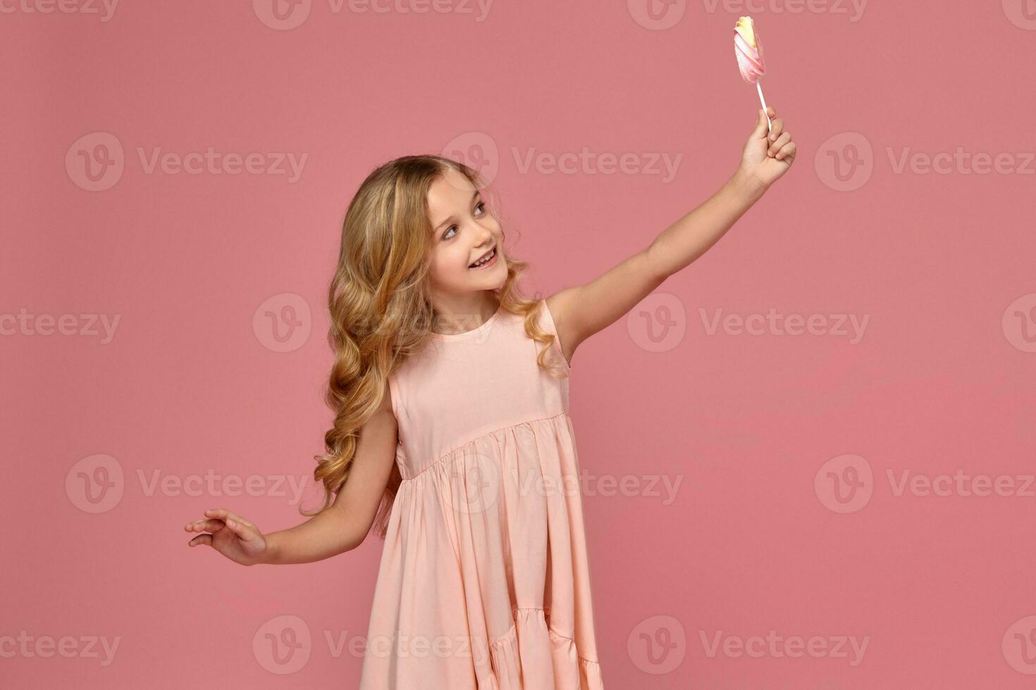 Little girl with a blond curly hair, in a pink dress is posing with a candy photo