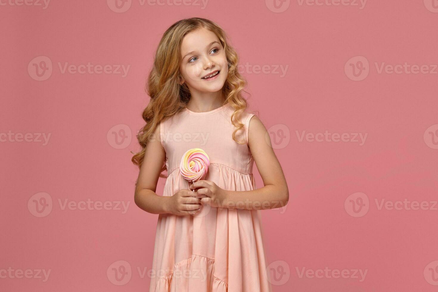 Little girl with a blond curly hair, in a pink dress is posing with a candy photo