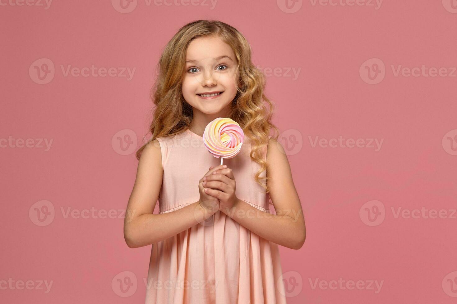 Little girl with a blond curly hair, in a pink dress is posing with a candy photo
