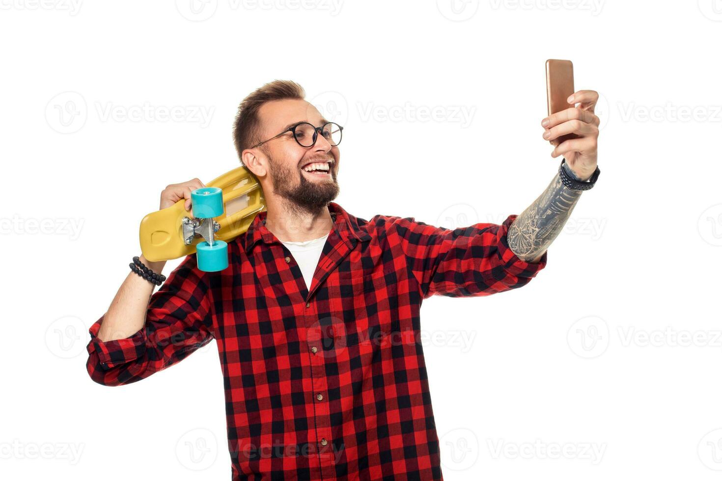 Young man holding the skateboard on shoulder raising his phone takes a selfie on a white background. photo