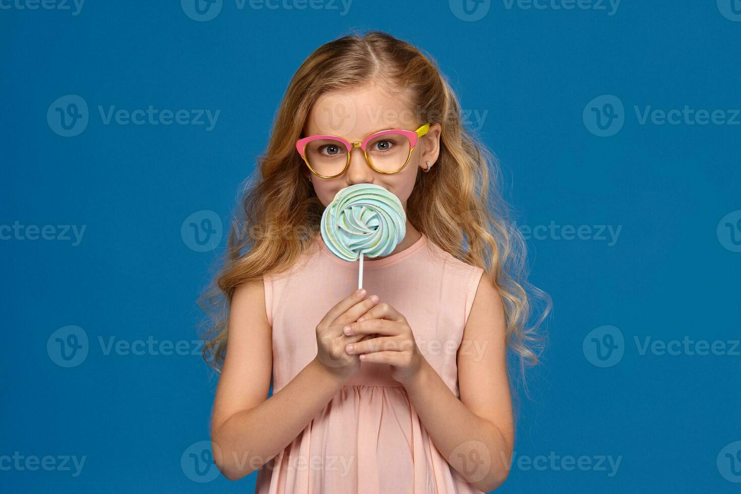 Fashionable little girl in a pink dress and glasses, with a candy in her hands, standing on a blue background. photo