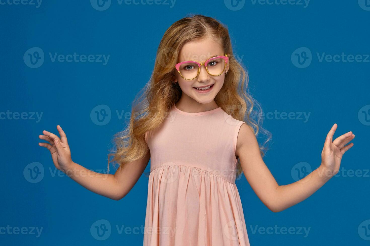 Fashionable little girl in a pink dress and glasses is standing on a blue background. photo