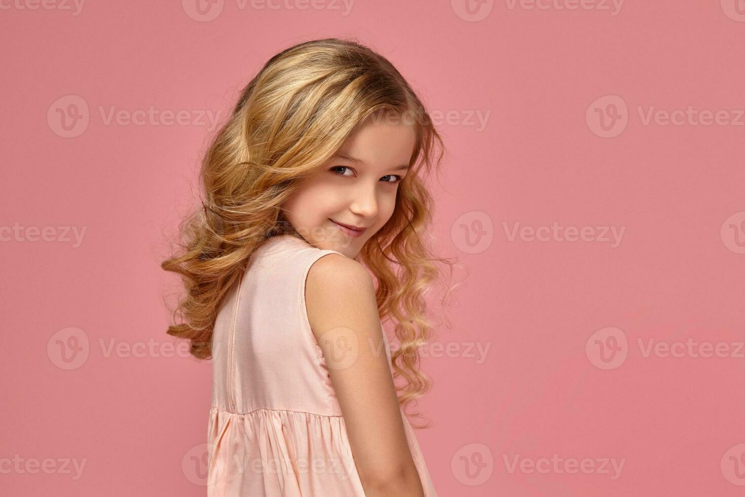 Little girl with a blond curly hair, in a pink dress is posing for the camera photo