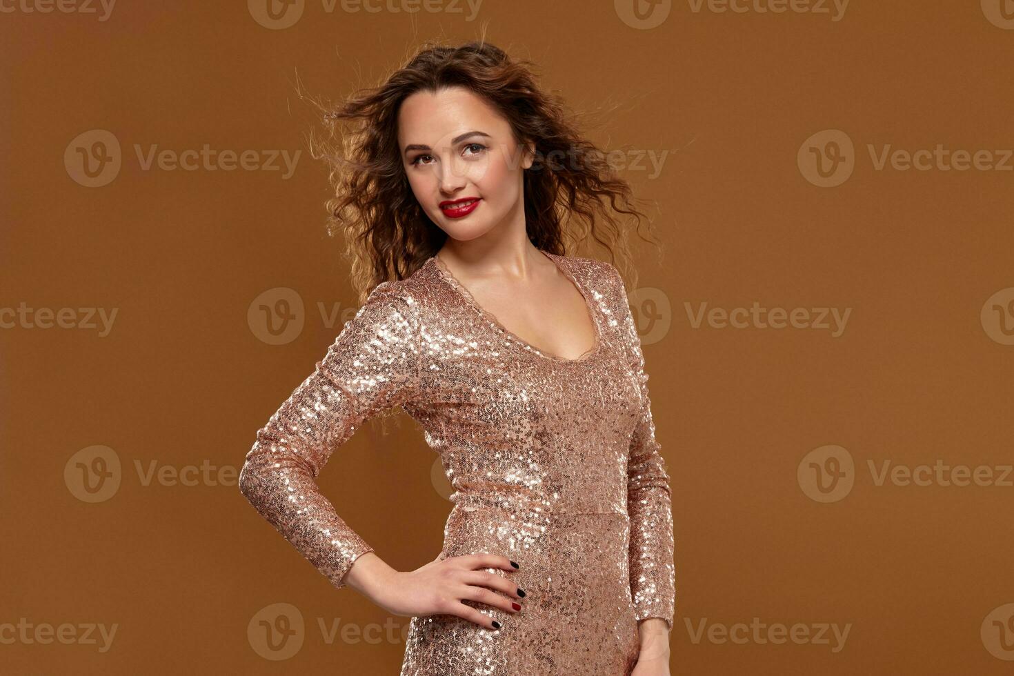 Portrait of young caucasian female in golden cocktail dress looking into the camera on brown background photo
