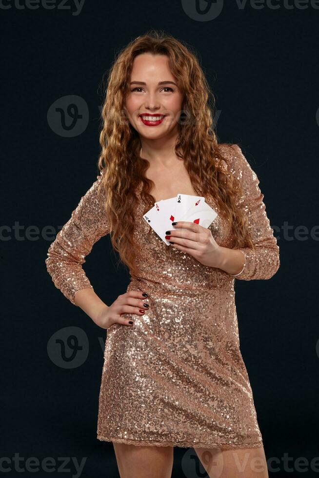 Beautiful young emotional woman in cocktail dress showing her cards to opponent, four of aces, best hand. Blcak background, studio shot photo