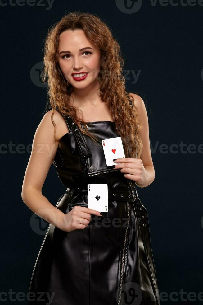 Emotional pretty young redhead woman holding pair of aces in black leather dress on black background photo