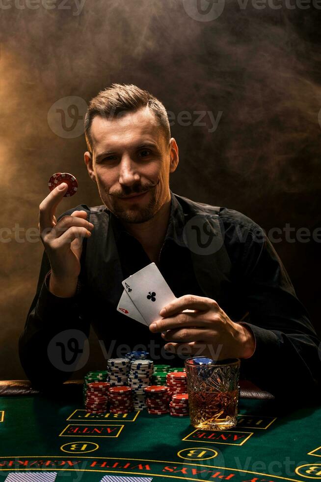 hombre es jugando póker con un cigarro y un whisky, un hombre espectáculo dos tarjetas en el mano, victorioso todas el papas fritas en el mesa con grueso cigarrillo fumar. foto