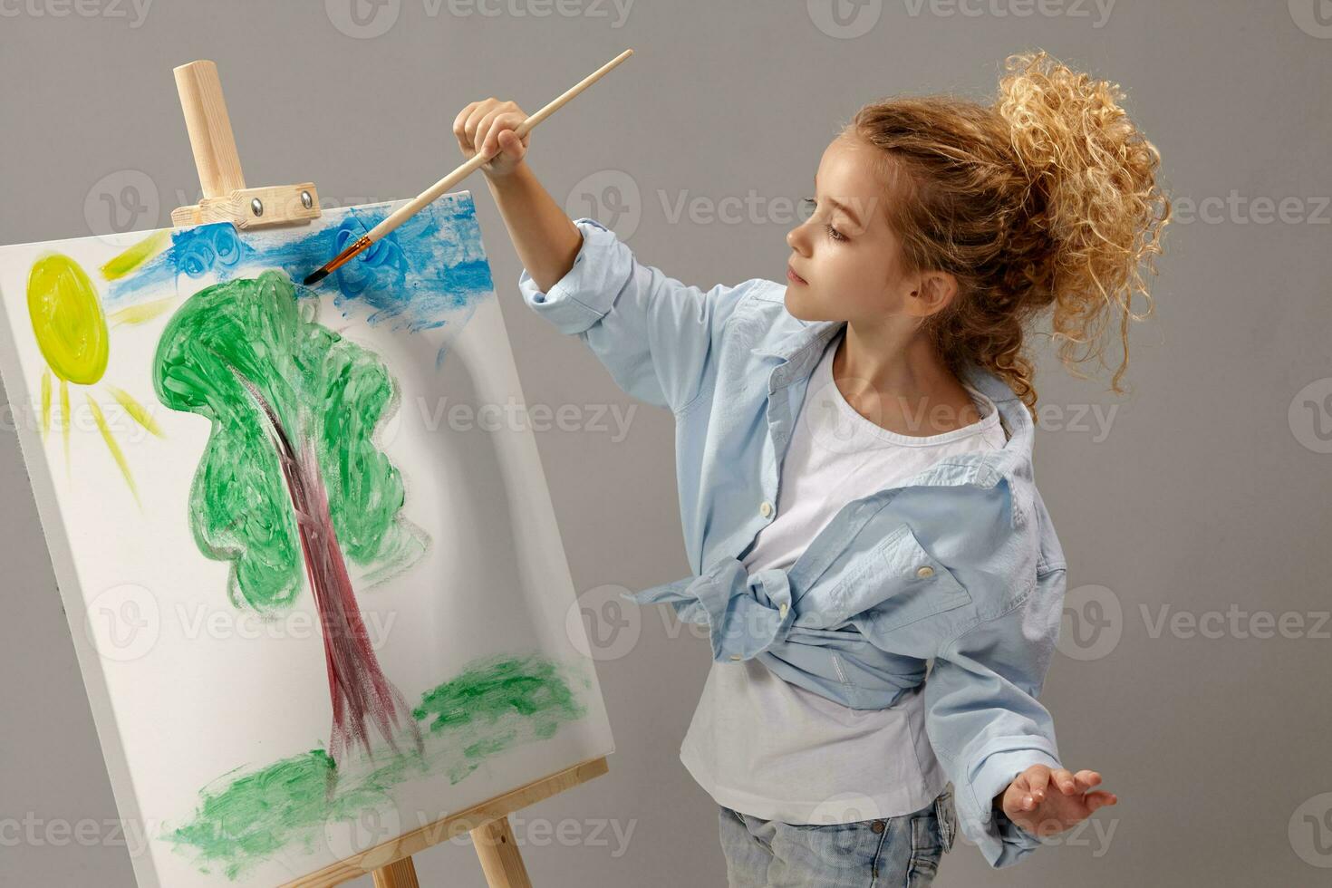 encantador colegio niña es pintura con un acuarela cepillo en un caballete, en pie en un gris antecedentes. foto