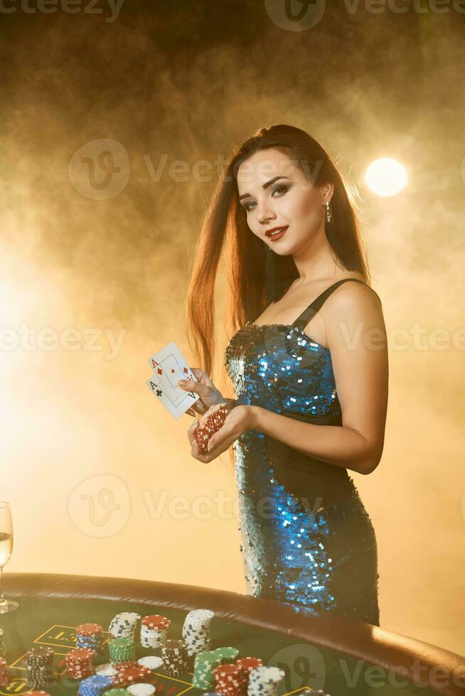 Young beautiful woman poses near poker table in luxury casino. Passion, cards, chips, alcohol, dice, gambling, casino - it is as female entertainment. Smoke background. photo