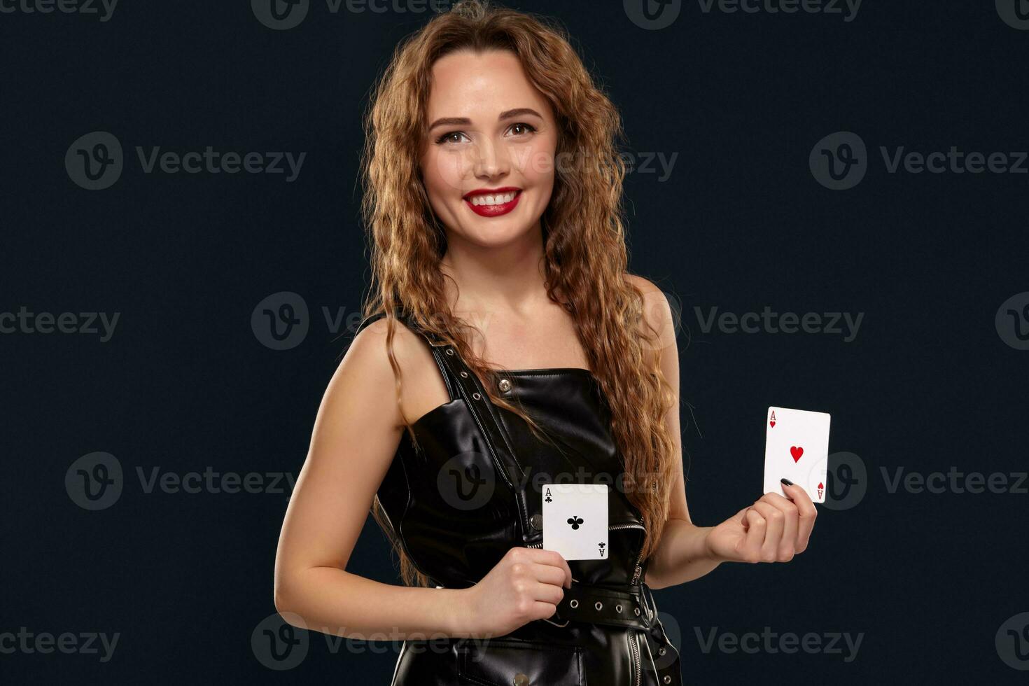 Emotional pretty young redhead woman holding pair of aces in black leather dress on black background photo