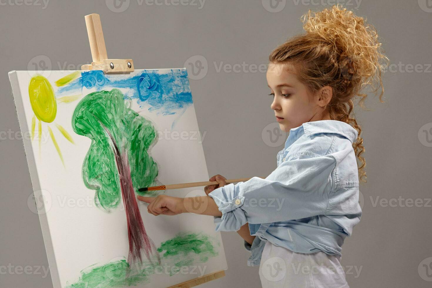 Charming school girl is painting with a watercolor brush on an easel, standing on a gray background. photo