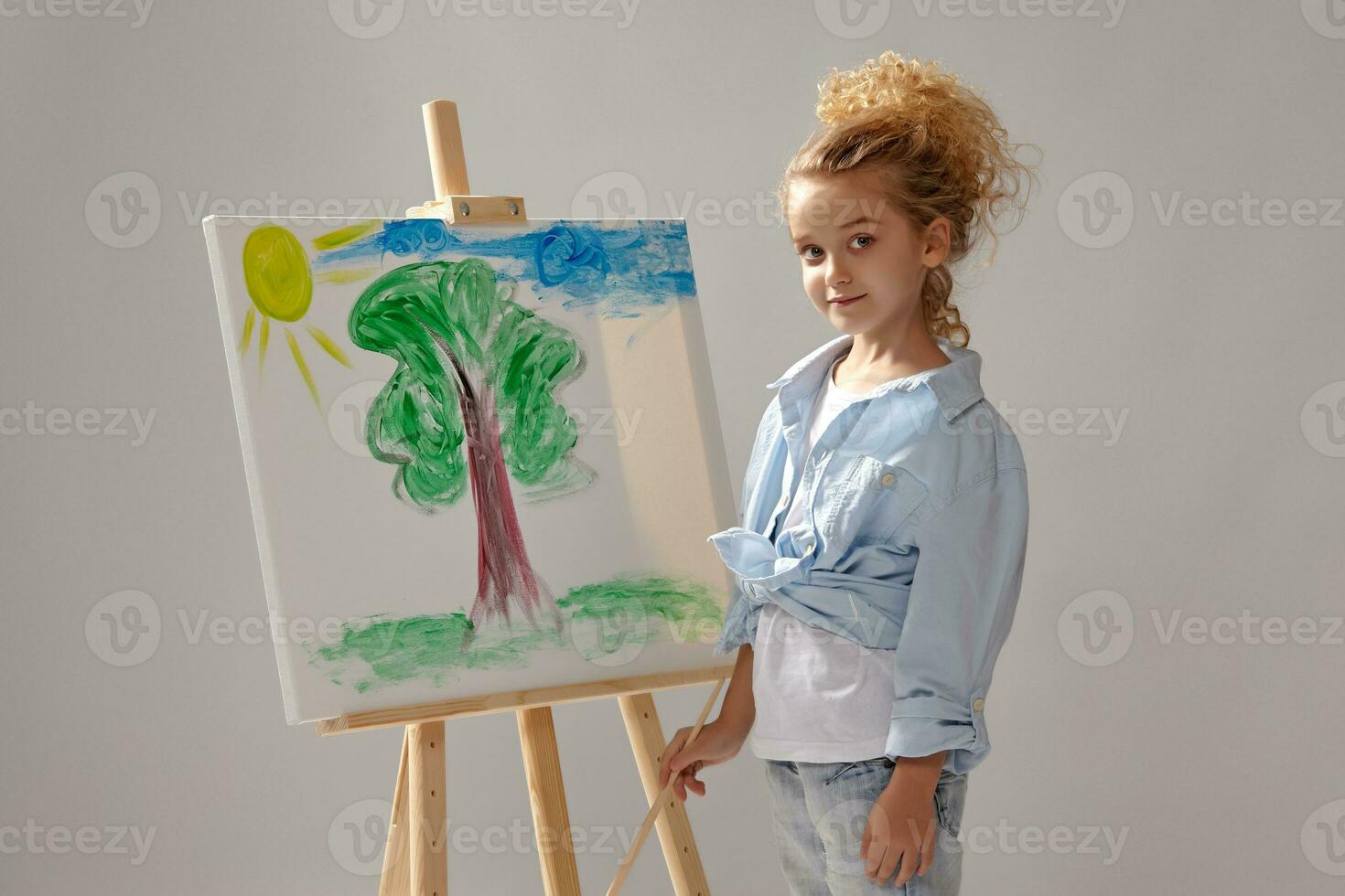 Charming school girl is painting with a watercolor brush on an easel, standing on a gray background. photo