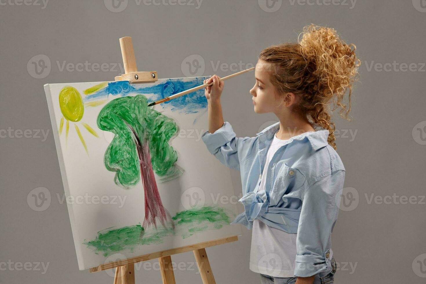 Charming school girl is painting with a watercolor brush on an easel, standing on a gray background. photo