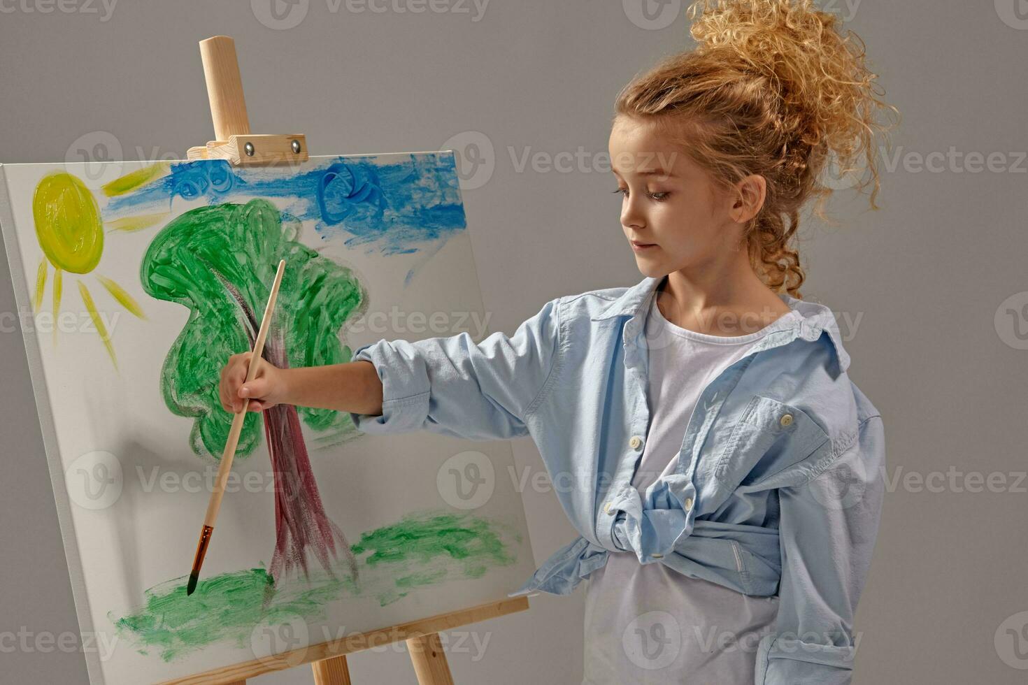 Charming school girl is painting with a watercolor brush on an easel, standing on a gray background. photo