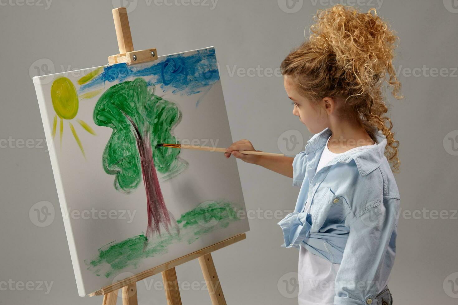 Charming school girl is painting with a watercolor brush on an easel, standing on a gray background. photo