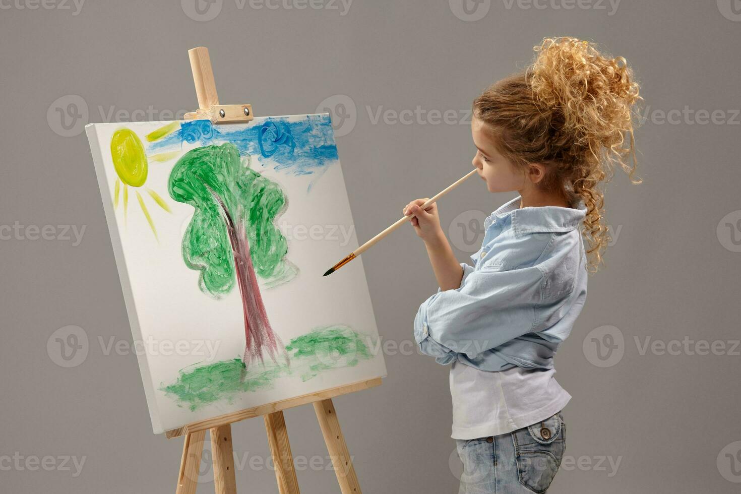 Charming school girl is painting with a watercolor brush on an easel, standing on a gray background. photo