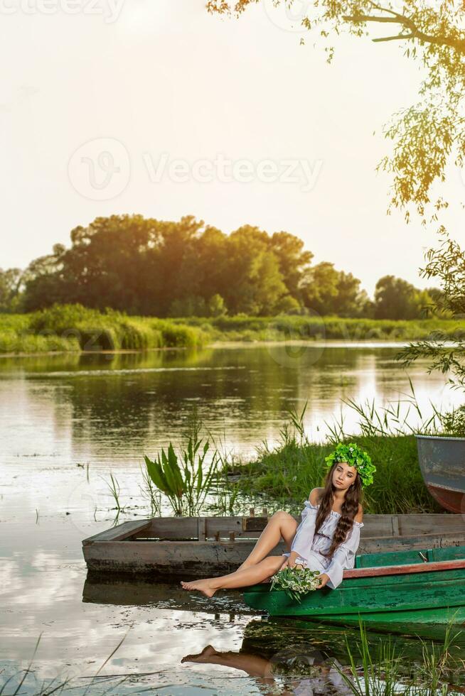 Young woman with flower wreath on her head, relaxing on boat on river at sunset. Concept of female beauty, rest in the village photo