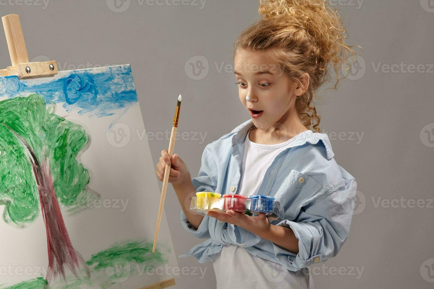 Charming school girl is painting with a watercolor brush on an easel, standing on a gray background. photo