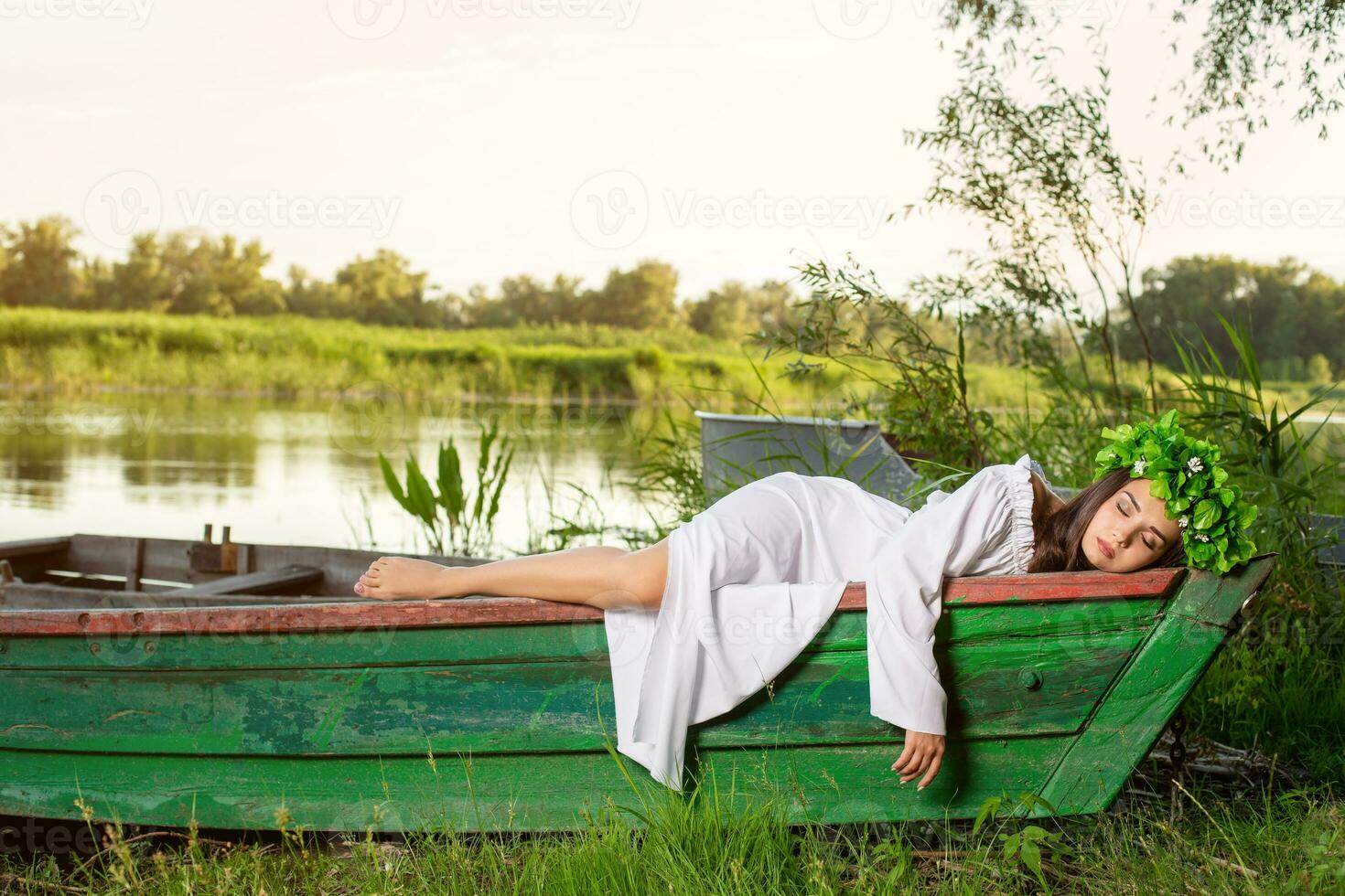 The nymph with long dark hair in a white vintage dress sitting in a boat in the middle of the river. photo