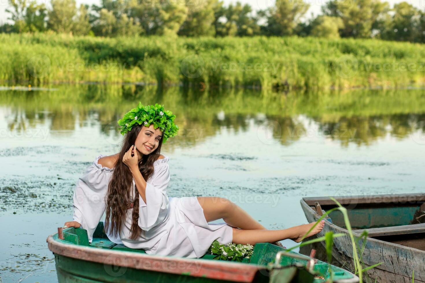 The nymph with long dark hair in a white vintage dress sitting in a boat in the middle of the river. photo
