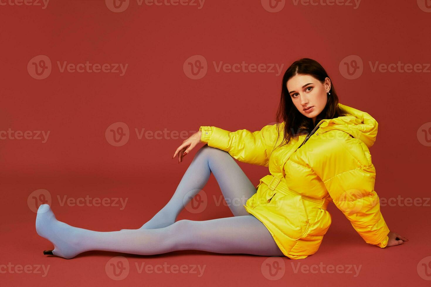 Studio portrait of young brunette woman sitting on the floor in yellow down jacket and grey blue panty hoses or stockings photo