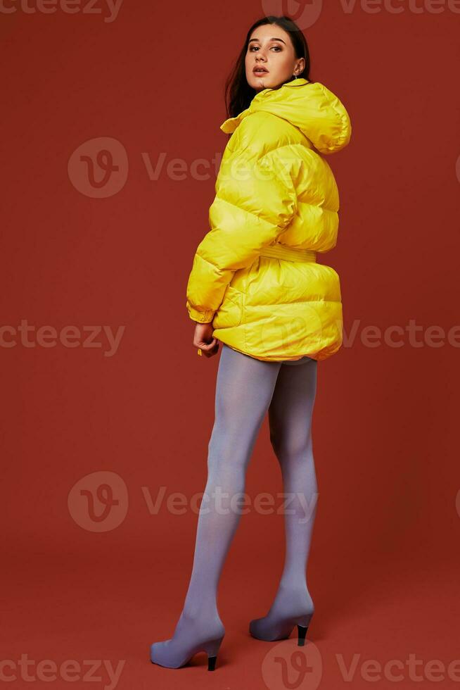 Studio portrait of young brunette woman in yellow down jacket and grey blue panty hoses or stockings. Studio shot photo