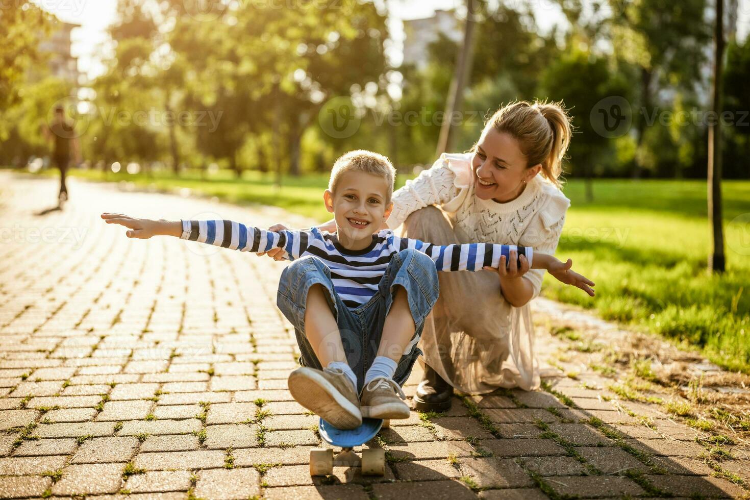 Mother having fun with her son in park on sunny day. Boy is ridding skateboard and his mother is pushing him. photo