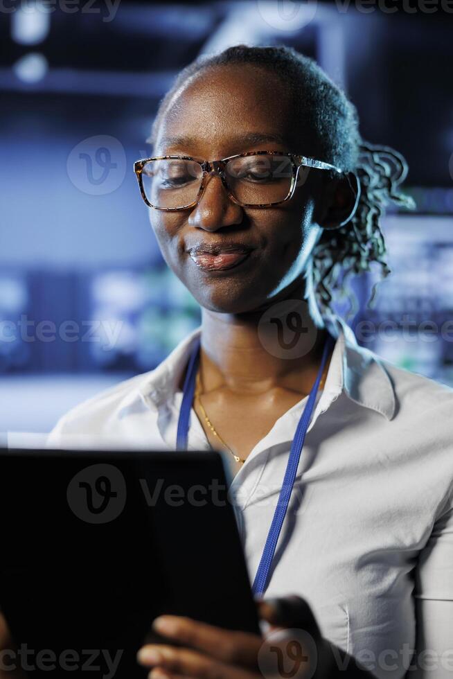 Satisfied admin in data center uses tablet to prevent system electronics overload. Efficient expert between server mainframes ensuring enough network bandwidth for smooth operations photo