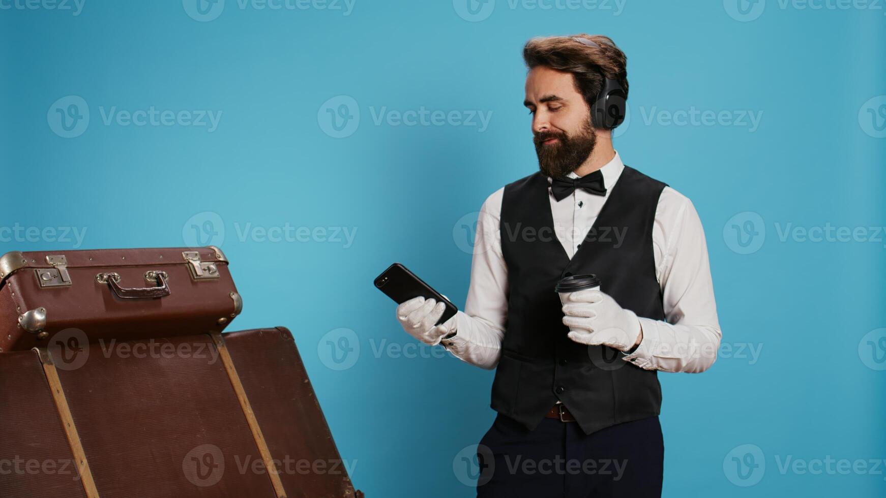 Modern doorman texting and drinking coffee in studio, enjoying work break with music on headphones. Young bellboy with professional hospitality industry job using phone and serving cold brew. photo