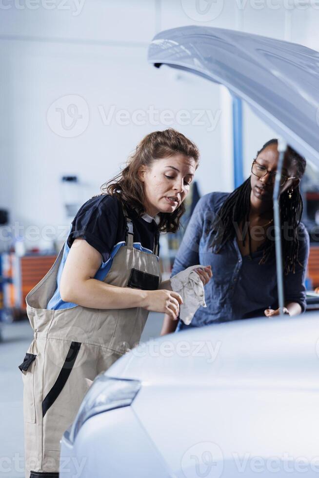 Engineer in garage finishing fixing car for BIPOC woman, looking underneath vehicle hood to remove remaining oil leaks. Worker does routine exhaust system cleaning on client automobile photo