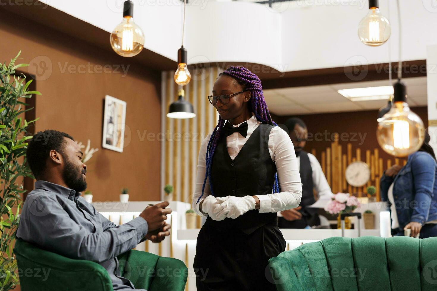 Smiling friendly African American woman hotel employee in uniform talking and interacting with happy guest in lobby. Generous rich man traveler giving cash tip to waitress girl at resort photo