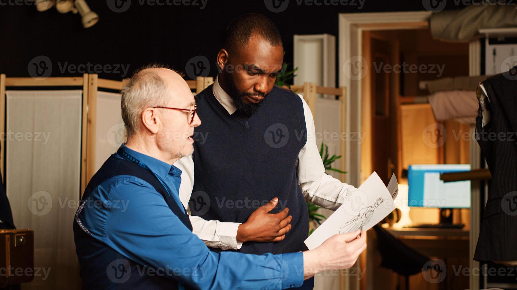 Designer and trainee reviewing drawings in atelier workshop, learning to create custom made clothes. Experienced tailor and student discussing about design and fabric. Handheld shot. photo