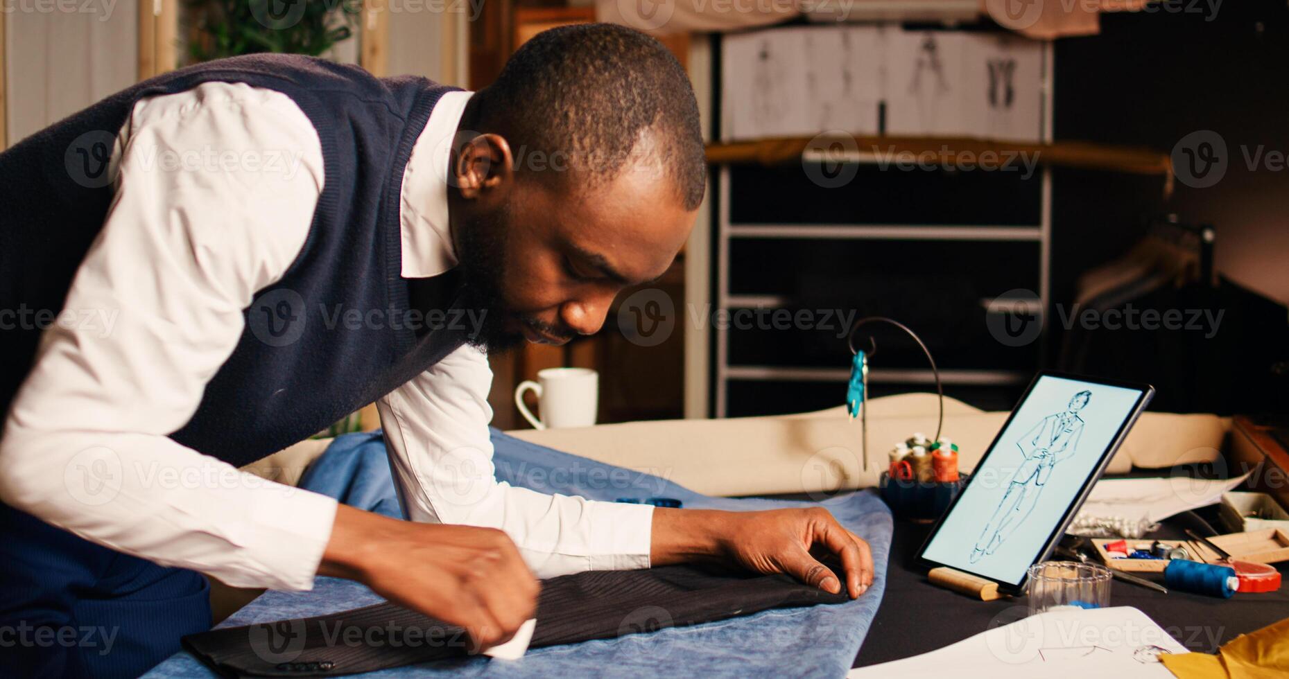 Male couturier marking fabric design to craft fashion items in tailoring atelier, using chalk to measure garment for needlework. Designer working on clothing line creation, workshop. photo