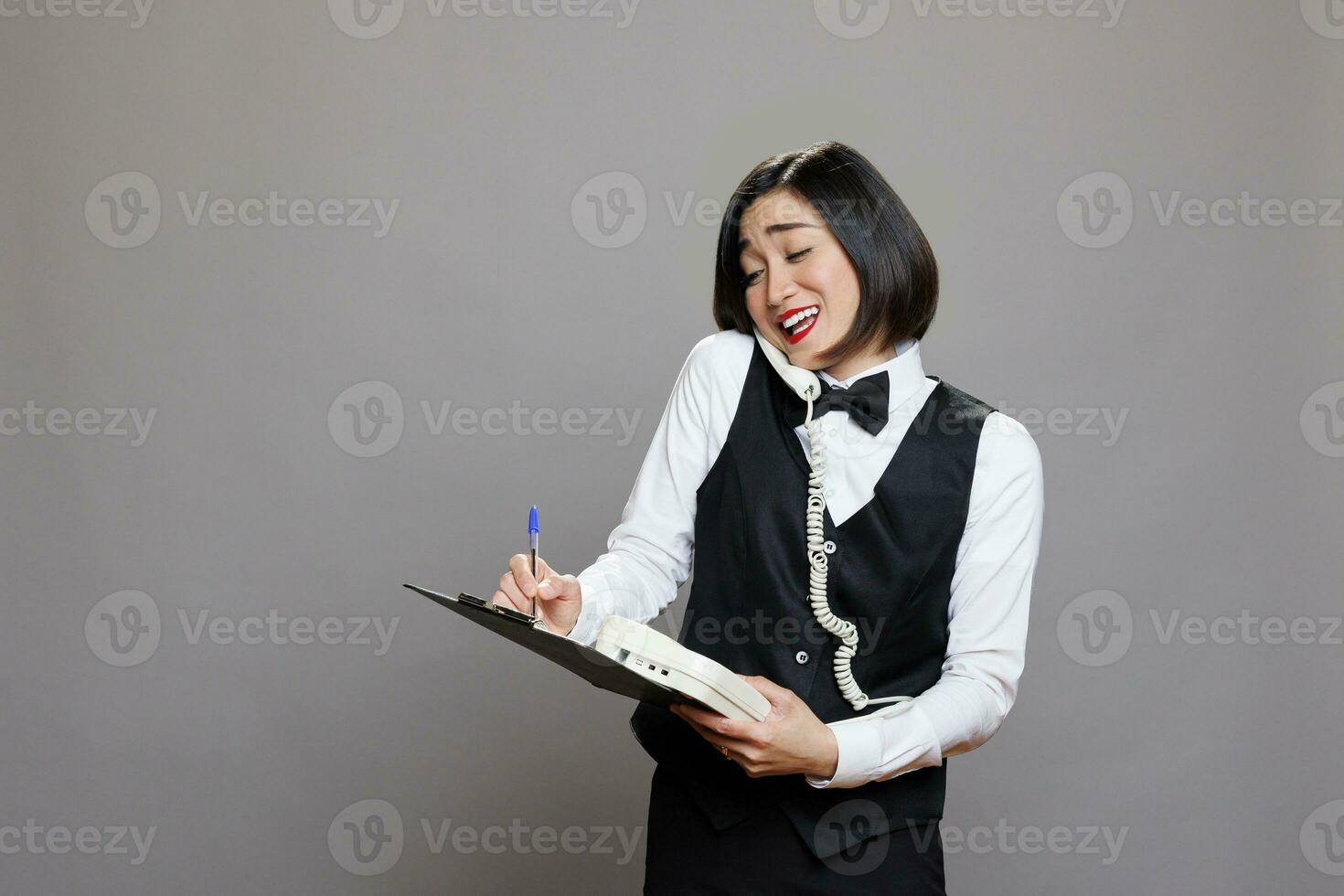 Smiling asian receptionist chatting with client on landline phone and writing in clipboard. Young attractive waitress in uniform having friendly conversation on retro telephone photo