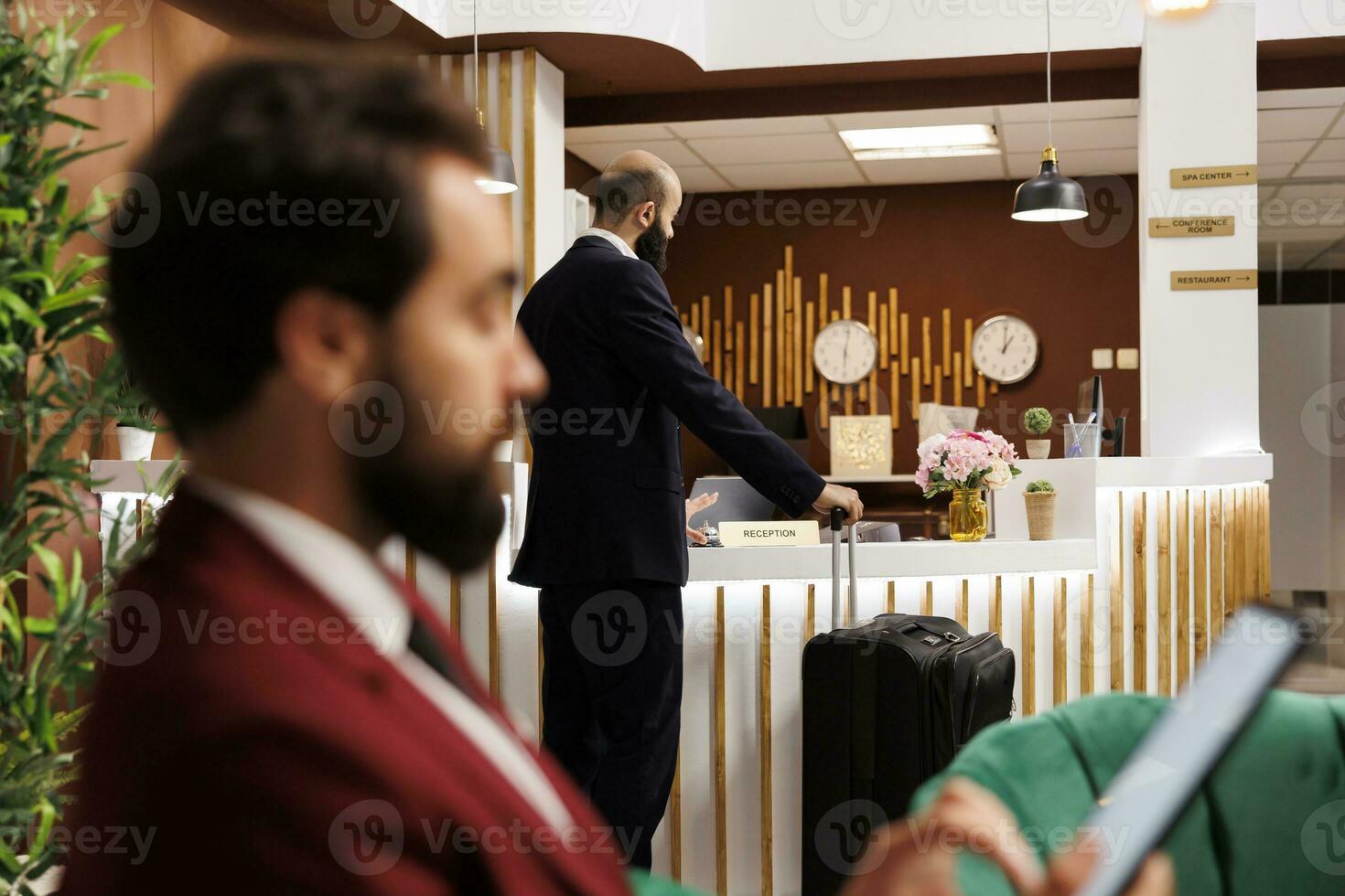 Businessman with luggage at front desk waiting to see room reservation and check in, travelling for work. Company professional preparing for upcoming business conferences, hotel lobby. photo