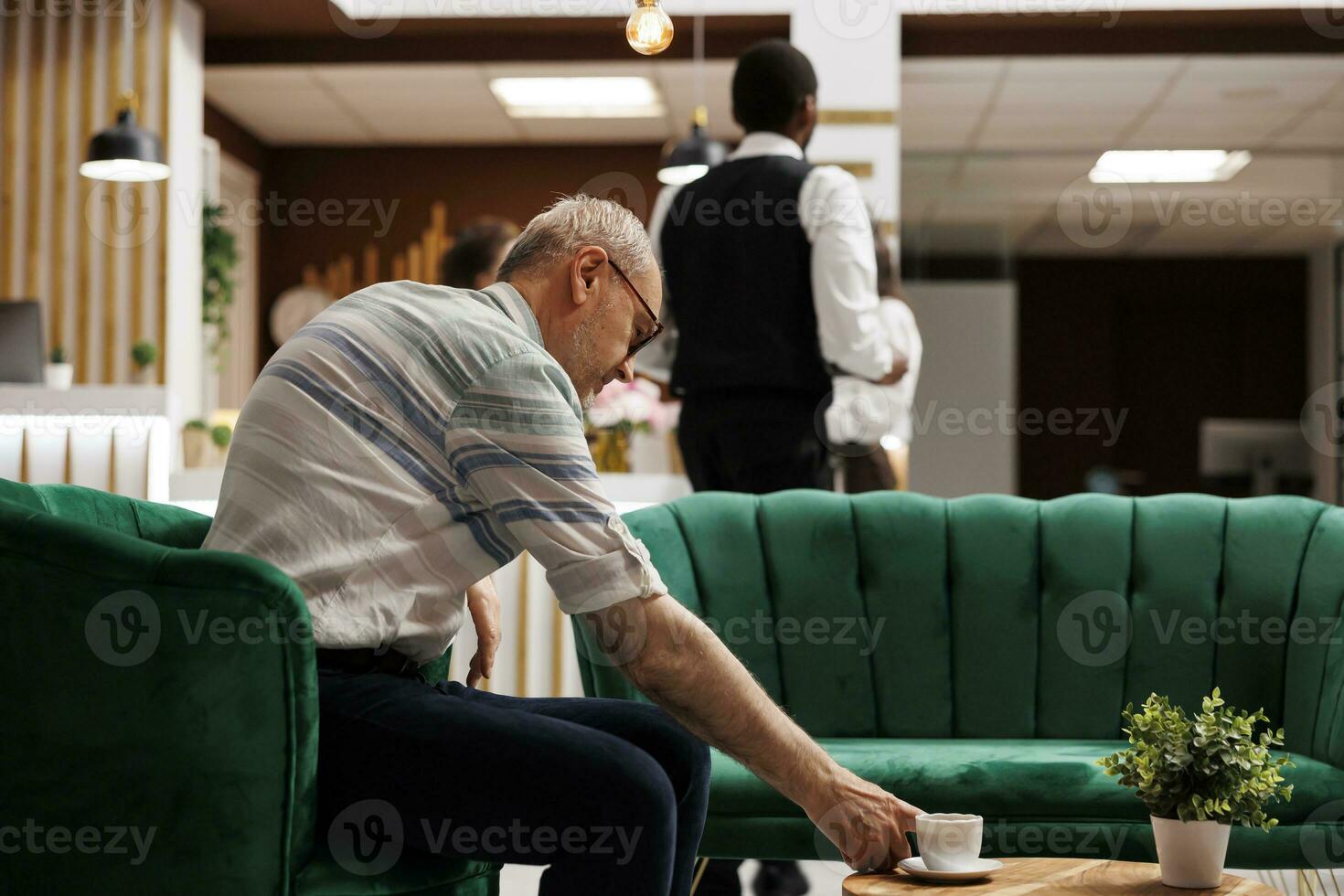 At elegant lounge area for checkin, elderly caucasian man sits on sofa reaching for cup of coffee on table. Senior guy in hotel lobby, waiting on the couch for the booking procedure. photo
