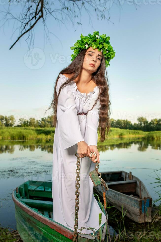 Young woman with flower wreath on her head, relaxing on boat on river at sunset. Concept of female beauty, rest in the village photo