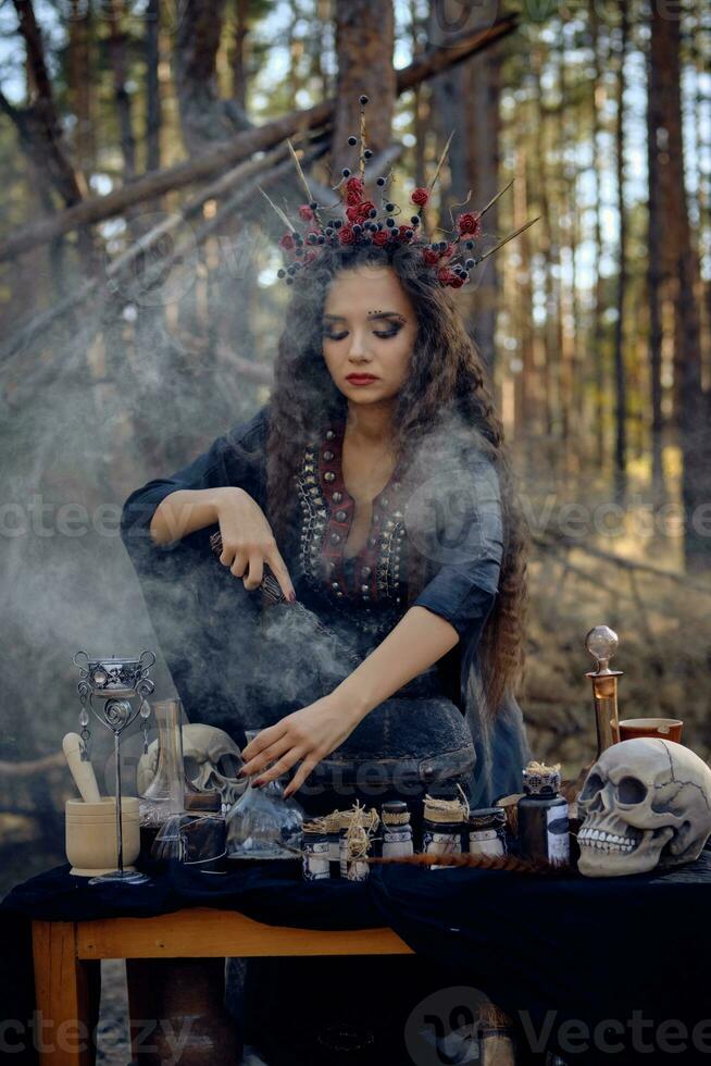 Witch in black, long dress, with red crown in her long hair. Posing in pine forest. Making a magic potion. Spells and witchcraft. Close-up, smoke. photo