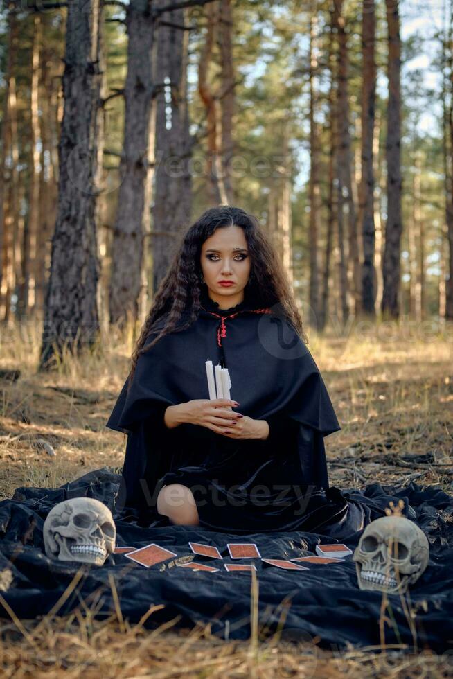 Witch in black dress with cape and hood. Posing in pine forest. Sitting on dark blanket whith fortune-telling cards and skulls on it. Full length. photo