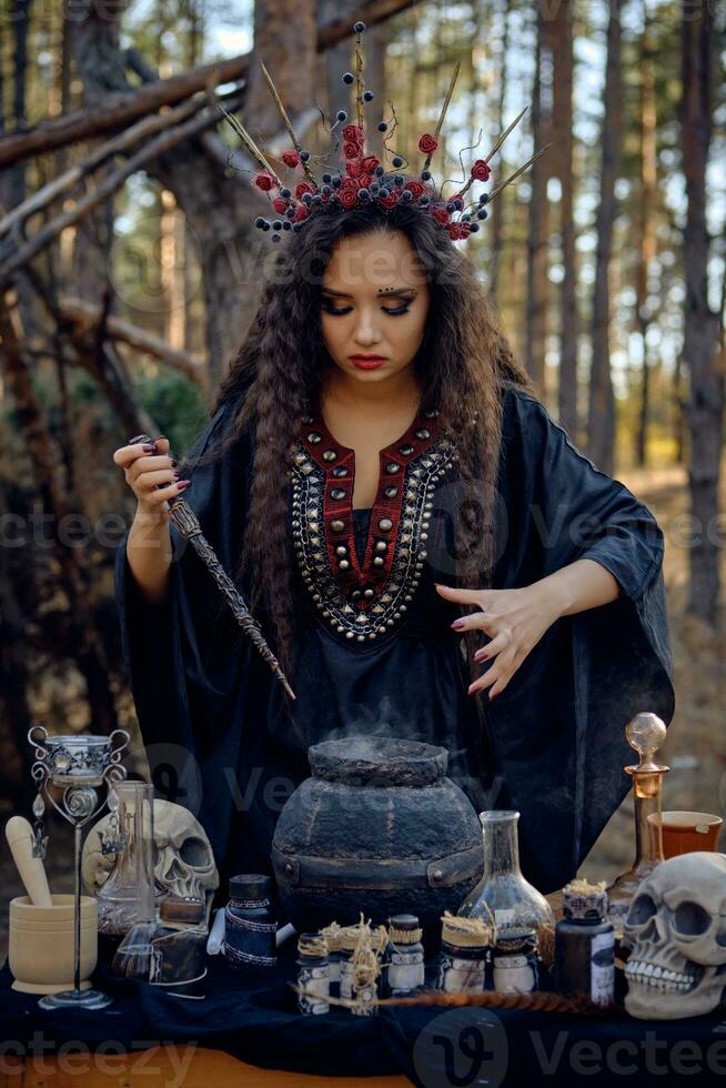 Witch in black, long dress, with red crown in her long hair. Posing in pine forest. Making a magic potion. Spells and witchcraft. Close-up, smoke. photo